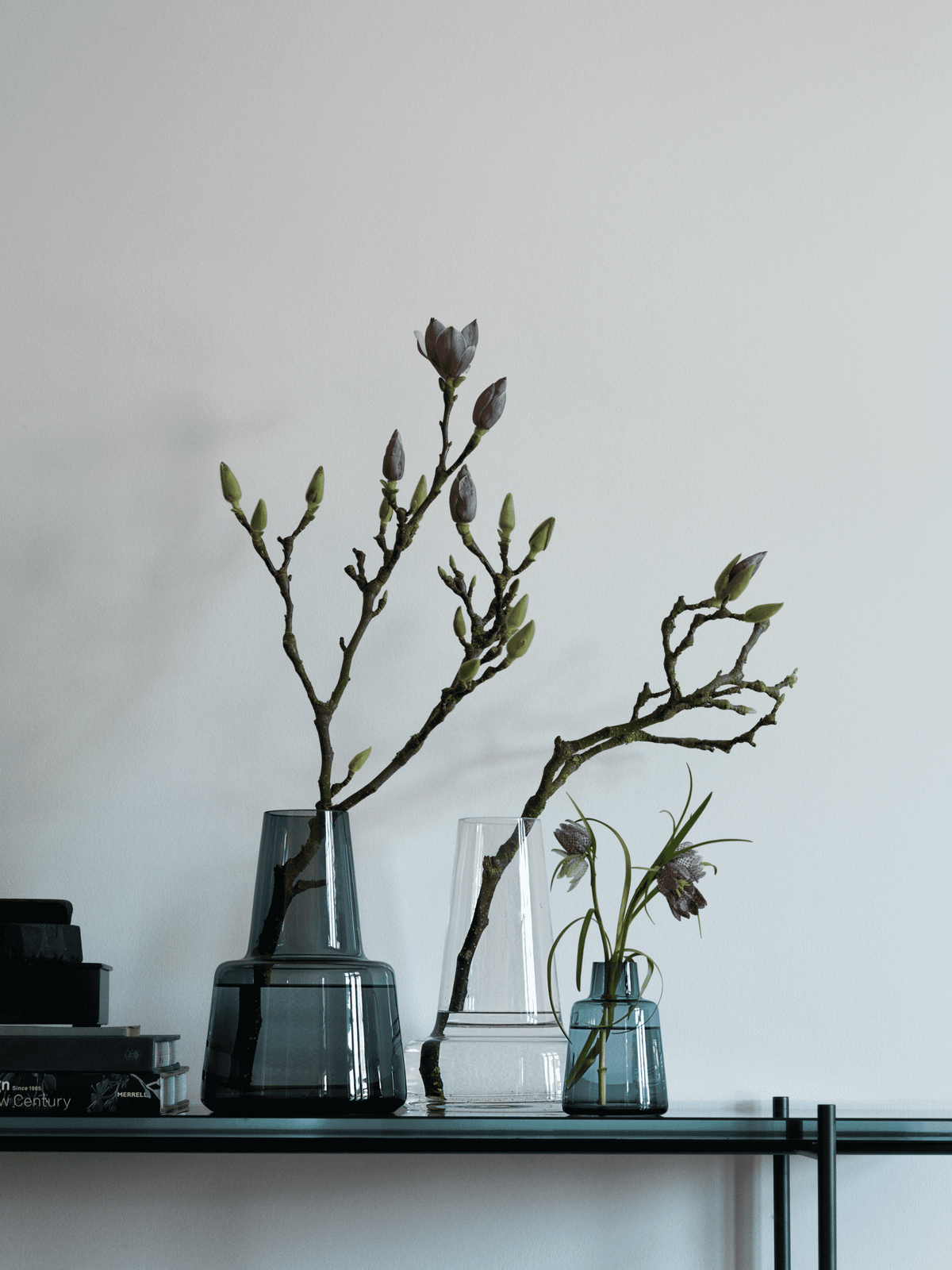 Holmegaard Flora medium neck vase in smoke glass with budding magnolia branches on a console, beside smaller Flora vases