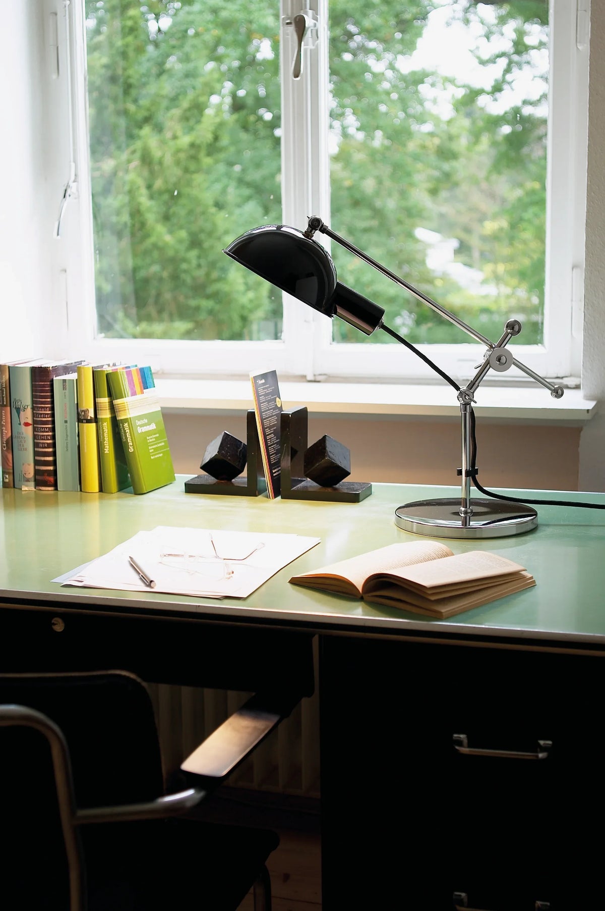 SF 27 desk lamp with chrome joints and black reflector on a green desk by a window, with books and an open notebook