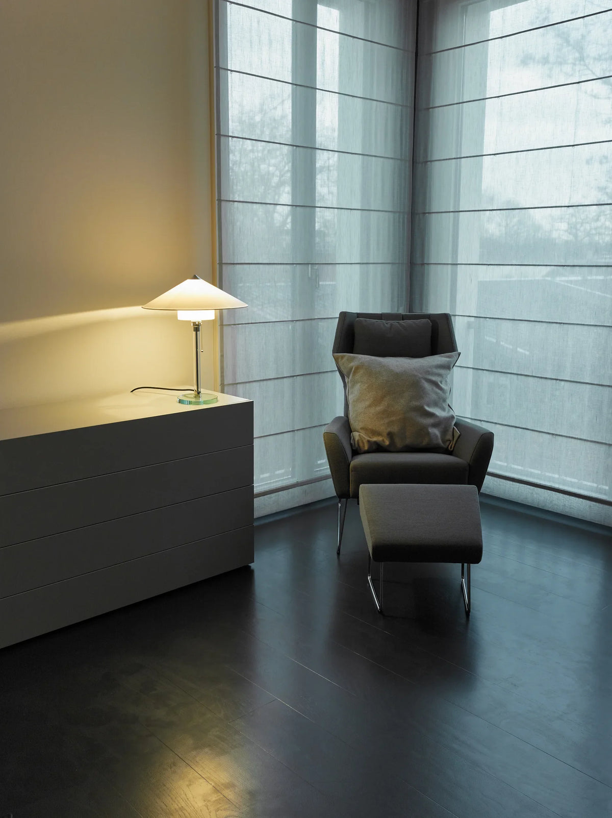 WG 27 Wagenfeld table lamp lit on a low sideboard beside a grey armchair and footstool in a corner with tall windows