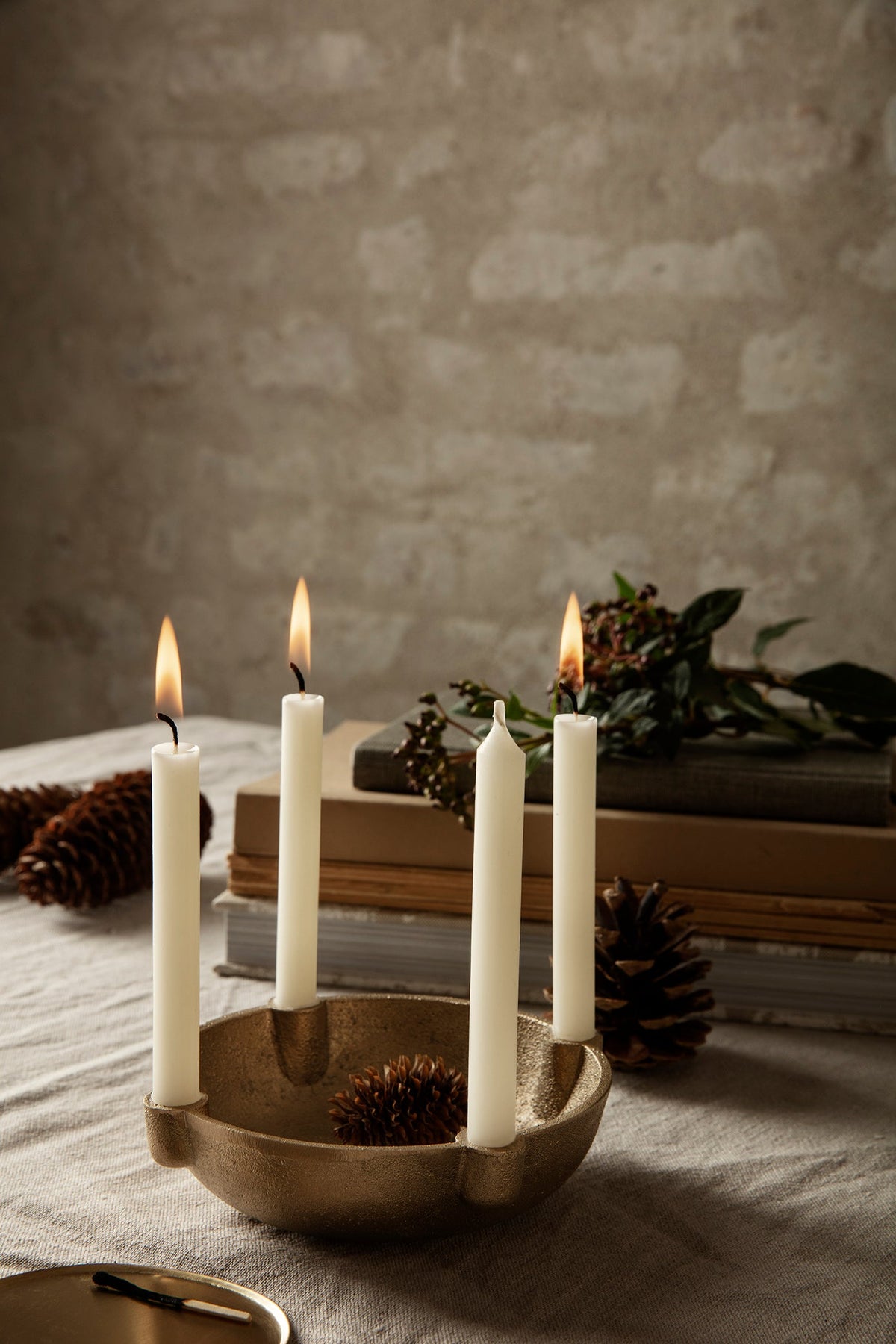 ferm LIVING Bowl Candle Holder Small in cast brass with four lit white candles on a linen tablecloth with pine cones