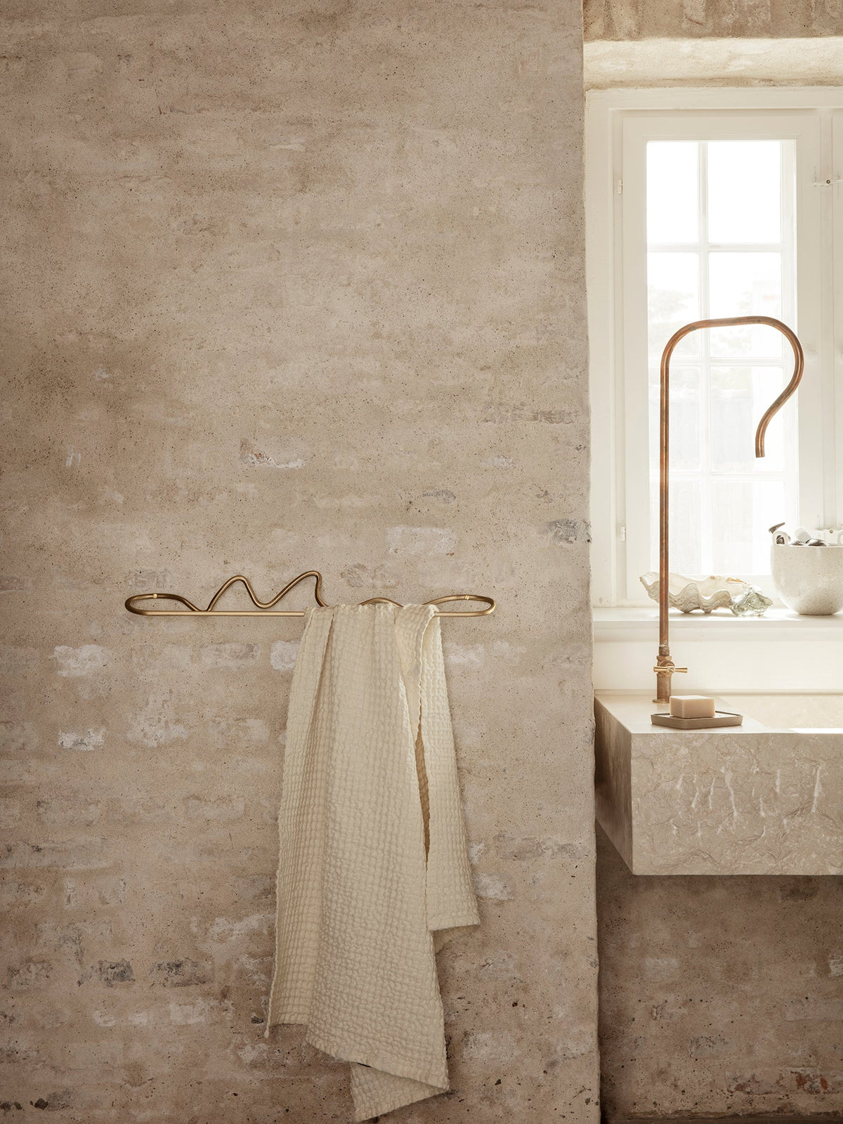Curvature towel hanger in brass with a cream towel on a plaster wall beside a stone basin and brass tap